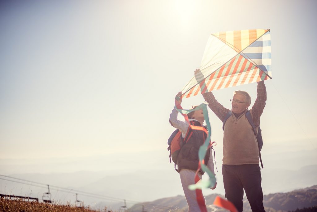 Grandfather flying a kite with his grandson on mountain.They wear casual clothes and enjoy in great sunny day in nature.