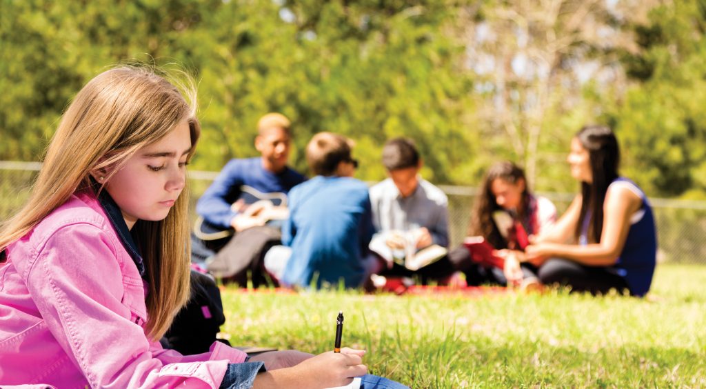 Pretty, blond pre-teen girl studies, writes notes at a local park. Open notebook. Friends in background study, listen to music.